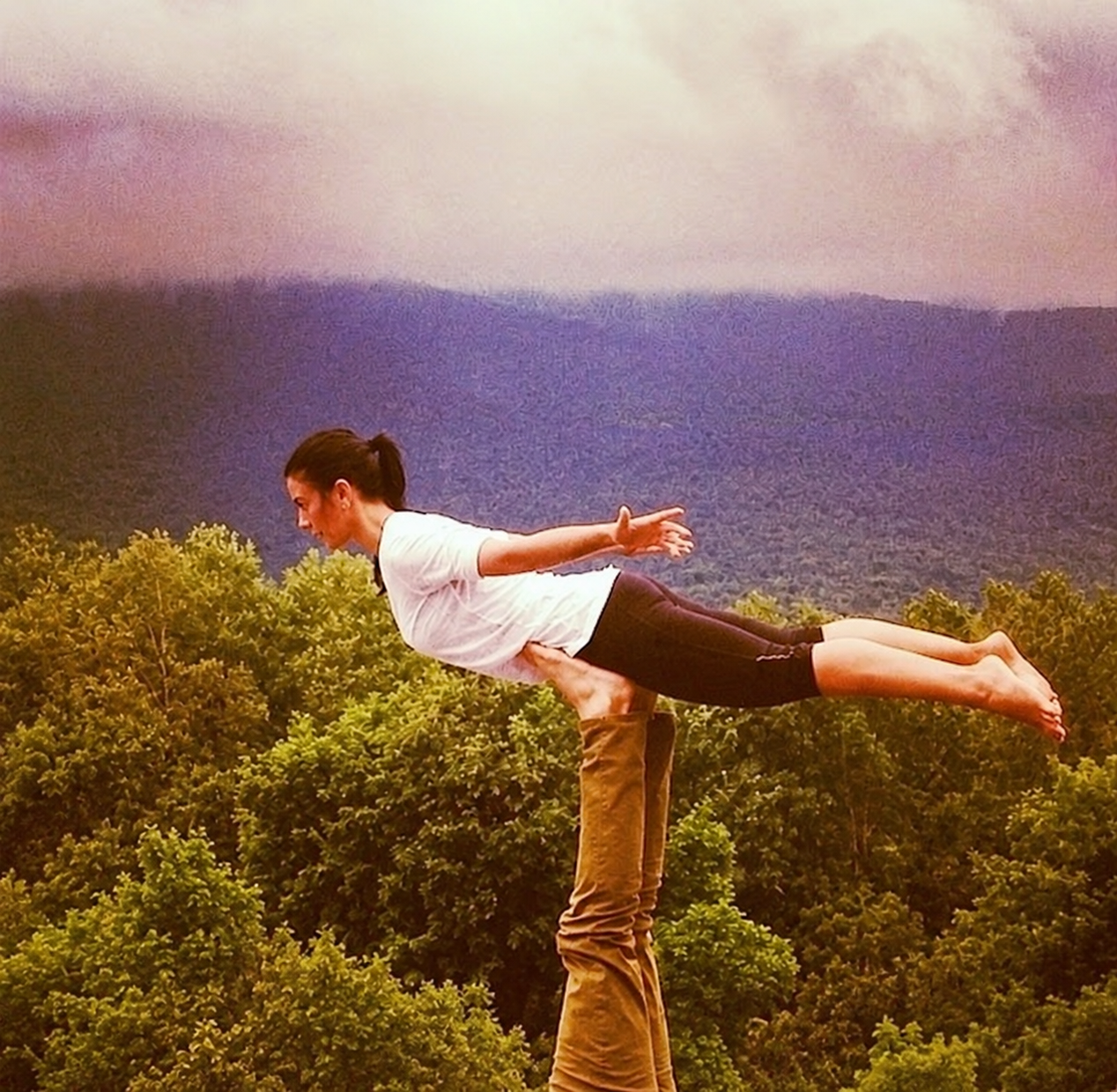 Georgia Zafferes balanced in an acro-yoga flying pose above a misty, forested mountain valley